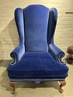 Front view of antique style blue velvet wingback chair showing brass studs on arms and wings, plush seat cushion, and carved wooden legs.