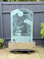 Front view of tall glass art deco style sculpture with frosted floral and geometric design, mounted on chrome base, shown outdoors against a fence.