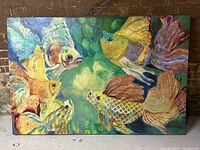 Full front view of the colorful fish canvas painting leaning against a brick wall on the floor.