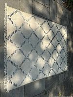 Full view of a rectangular flat woven area rug laid on outdoor stone tiles, showing a cream base with a black and grey geometric diamond lattice pattern with fringed edges on one short side.