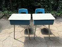 Two blue metal classroom desks with light-colored desktops and attached blue molded plastic chairs shown from the front, placed outdoors on cracked concrete.