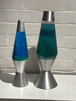 Two lava lamps side by side against a brick wall showing size difference and blue-green liquids