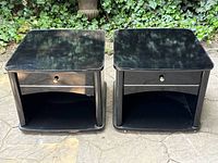 Pair of black high gloss nightstands shown outdoors on stone surface with ivy background.