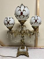 Front view of vintage Hollywood Regency table lamp with three frosted glass globes decorated with gold vine motifs and red gem accents, ornate cherub figure, crystal prisms, and white marble base.
