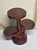 Front angle photo of the wooden plant stand showing three round platforms at different heights with natural wood grain and knots visible. Moderate surface wear and chips shown.