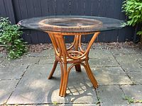 Round vintage table with clear circular glass top and rattan base shown outdoors on paved surface from side angle