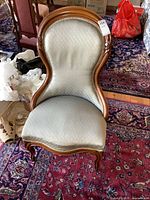 Front view of antique wooden frame upholstered chair showing curved back and seat, light beige upholstery, placed on patterned rugs with minor wear.