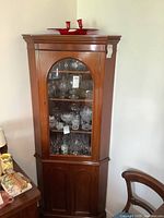 Full view of the antique corner cabinet showing the wood finish, glass door with display shelves, and wooden two-door lower cabinet.