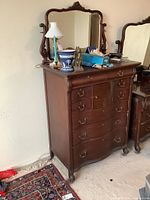 Full front view of the antique wooden high boy dresser with mirror, showing curved front and multiple drawers with brass handles, some objects on top.