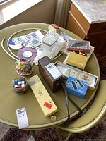 Photo showing vintage binoculars in brown case, Rubik's Cube, dominoes, glass coasters, floral porcelain centerpiece, wool winder box, assorted cards, and other small items spread on a table