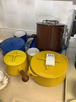 Six enameled pieces on countertop: brown stock pot, three yellow wooden-handled pans and two blue pans