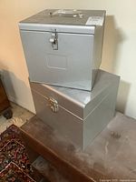 Pair of silver-colored metal file boxes stacked, showing size difference and metal clasp closures.
