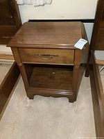 Front angled view of vintage wooden night stand showing single drawer with brass handle, open shelf below, and scalloped bottom apron.