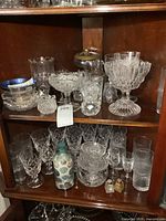 Two shelves of assorted clear glassware including candy dishes, stemware, pitcher, glasses, and decorative bottle.