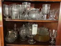 Full view of two wooden shelves with assorted glassware items including various glasses, a candy dish, vases, plates, and a pitcher.