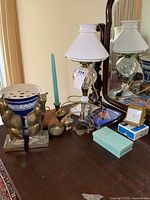 Wide shot showing vintage oil lamp converted to electric, brass cat bookends, Wedgwood planter, blue candle, small brass decor items, timer and wind-up clock on wooden table with mirror background.