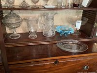 Shelf displaying all items in the lot: compote, pitchers, bread plate, trifle bowl, art glass footed bowl