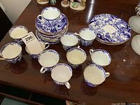 11 blue and white porcelain teacups and matching saucers stacked on a dark wooden table with 4 snack plates to the right.