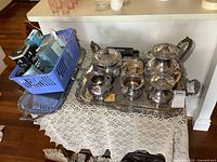 Silver plate tea and coffee set on a lace covered table, including pots, trays, and blue basket with polishing materials.