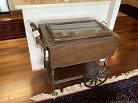 Vintage wooden tea wagon with glass top, drop leaf sides down, drawer, large wooden wheels, and caster wheels on hardwood floor next to rug.