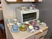 Wide view of all 16 teacup and saucer sets arranged on kitchen counter, showing variety of colors and styles.