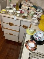 Full view of all 16 teacups and matching saucers arranged on drawers and stove area, showing variety of floral patterns and colors.
