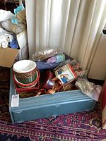 Vintage blue suitcase packed with woven baskets, decorative tins, potpourri, ribbons, and other assorted items on a patterned carpet.