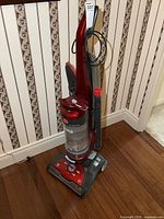 Full view of red Hoover upright vacuum with hose and wand, standing on a wooden floor near wallpapered wall.