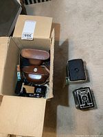 Box with multiple vintage cameras and accessories visible, including brown leather case