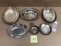 All six silver and silverplate pieces on cardboard with dollar bill for scale