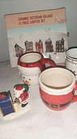 Full view of ceramic Victorian village lighting set box with two Santa-themed mugs and Santa figurine in front.