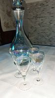 Iridescent glass decanter and four liqueur glasses arranged on a table, showing the height and iridescent finish of each item.