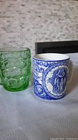 Photo of two mugs on a white cloth with a tiled background, showing the green depression glass beer mug and blue and white ceramic coffee mug with Gemini decoration.