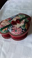 Heart-shaped jewellery box with colorful floral padded lid and small red bow on white surface.
