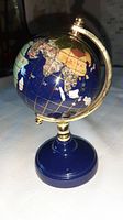 Miniature globe showing Africa, part of Europe, Asia with colorful inset stones over a blue ocean base. Globe mounted on blue round base with brass fittings and axis for rotation.