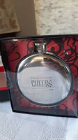 Front view of the unopened round stainless steel flask with embossed 'CHEERS' text inside packaging