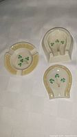 Three Belleek porcelain dishes consisting of one circular ashtray and two horseshoe-shaped dishes, all decorated with shamrocks and embossed pale yellow borders, displayed on a white textured fabric.