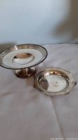 Photo showing both silverplated dishes on a white surface, cake stand on left, decorative plate on right.