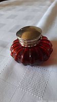Close-up photo of vintage cranberry glass inkwell with silver-plated top on white fabric surface, showing scalloped glass design and smooth lid with ridged edge.