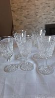 Five small wine glasses set on a white textured tablecloth, showing clear crystal with cut Lismore pattern.