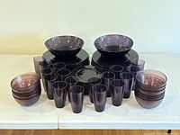 Full set of purple glass dishware arranged on table including stacked plates, bowls, and glasses.
