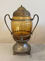 Front view showing amber glass barrel in metal holder with lid, handles, and spigot on front base.