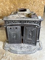 Front view of the cast iron stove with both decorative front doors open, showing ornate floral patterns and the stove base with Fawcett branding.