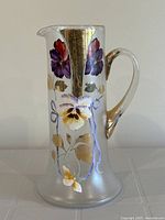 Front view of a tall clear blown glass pitcher with a curved handle, spout, and hand painted flowers and gold leaf decorations showing overall item.