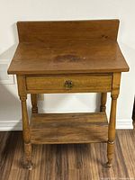 Front view of the antique pine washstand showing drawer, lower shelf, turned legs, and wood grain top with raised backsplash.