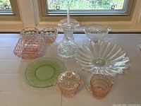 Overview of all glass pieces including pink bowls, dishes, green uranium plate, clear flower bowl, candlestick, and others on white table by window.