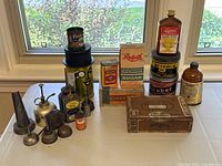 Full view of lot showing various antique tins, oil cans, bottles, sugar box, and cigar box arranged on white surface in front of window
