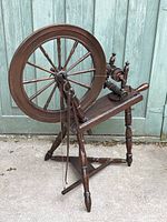 Side view of antique wooden spinning wheel showing large drive wheel and base structure, outdoors on concrete patio in front of teal wooden door.