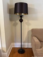 Front view of the entire lamp including black drum lampshade, tortoiseshell glass section, slender black metal pole, and round black metal base. Positioned beside a couch near a wall.