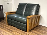 Front angled view of vintage reclining love seat showing oak frame and green leather upholstery on seats and backs.